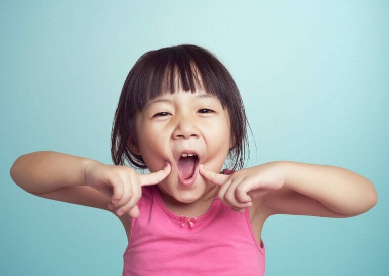 A young child in a pink shirt pulling the corners of their mouth outward