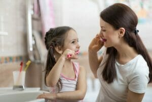 Parent and young child brushing their teeth together in a bathroom. Child brushing teeth with the recommended amount of fluoride toothpaste. How Much Toothpaste Should Your Child Use?