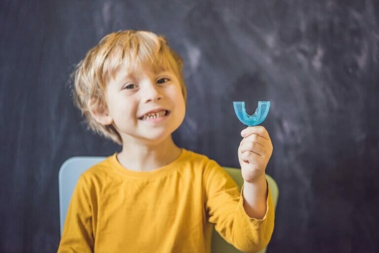 A young child in a yellow shirt smiling and holding up a blue dental retainer