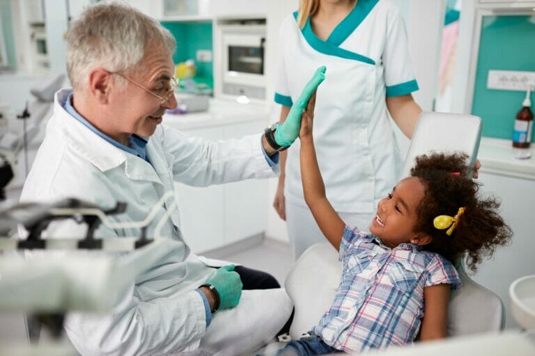 A dentist in a white coat high-fives a young girl in a dental chair